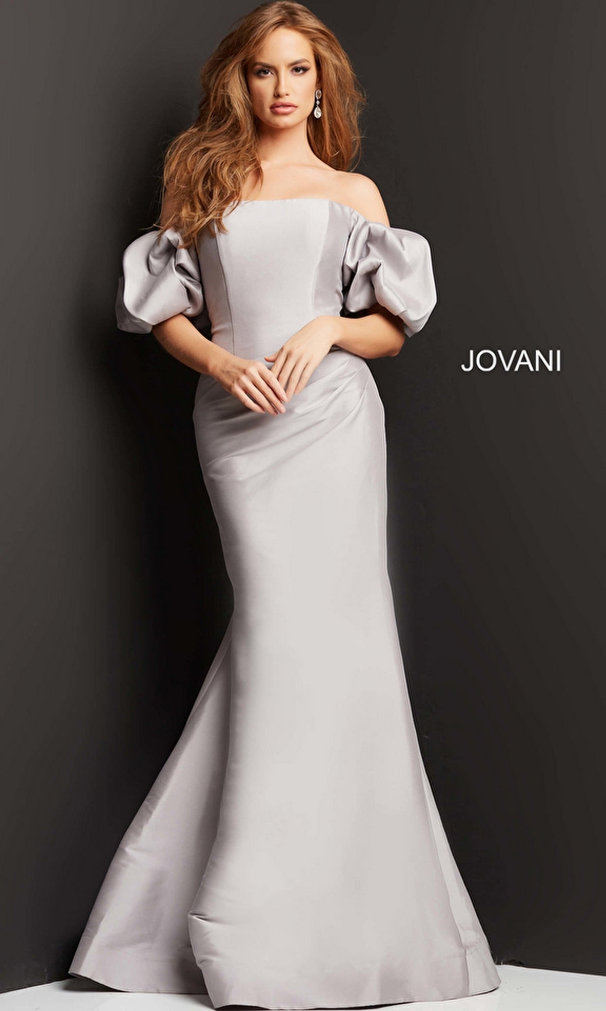 A woman poses in the Puff-Sleeve Mikado Evening Dress (Jovani 08361), a light gray off-the-shoulder gown with puffed sleeves and a mermaid silhouette, against a dark background.